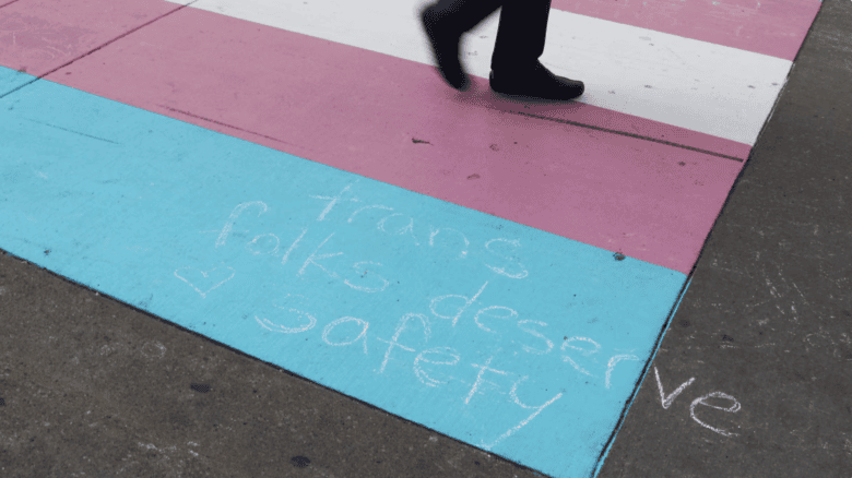 Feet are seen walking across a crosswalk painted in the colours of the trans flag. On the pavement written in what appears to be chalk are the words "Trans folks deserve safety."
